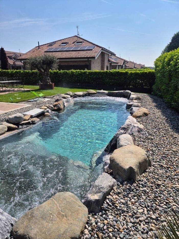 Piscina Rocks Design stretta e lunga tra massi e ciottoli, con un ulivo sullo sfondo